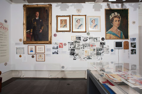 Wall oif the gallery featuring large dark portrait of man, three portraits of Queen Elizabeth,  photographs a, newspaper clippings and other ephemera